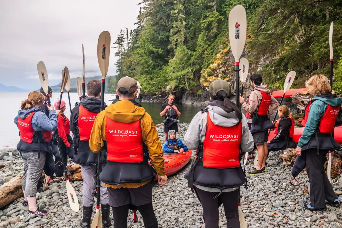 A guide briefs a group of guests on the pebble beach beside their sea kayaks.