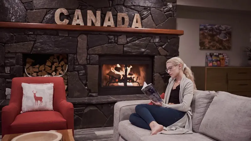 A guest reads on a sofa beside a stone fireplace under the word CANADA at a Basecamp property.