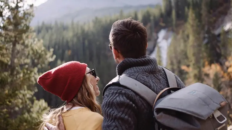 Two hikers in warm layers look out from a forested ridge toward a waterfall.