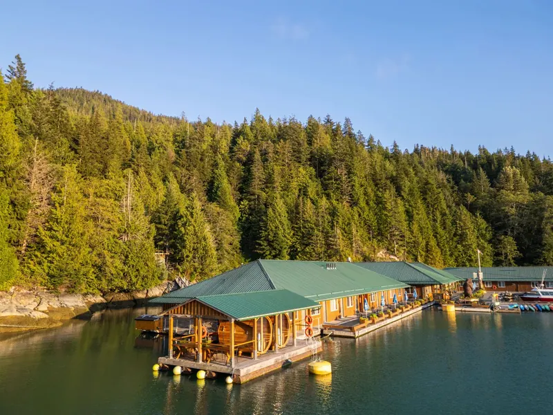 Evening sun lights the floating lodge’s green roofs along the shore of Knight Inlet below a forested hillside.