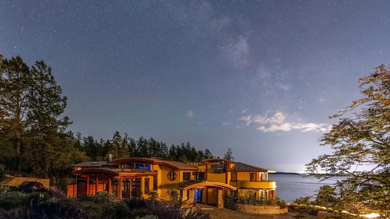 The house glows at night under a starry sky beside the water.