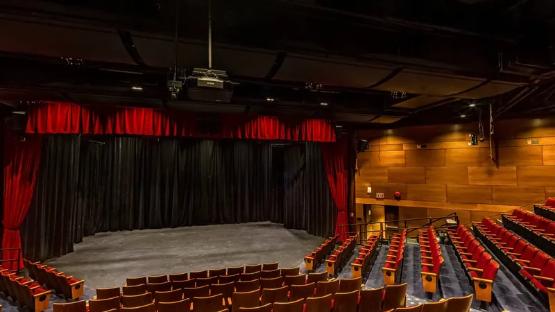 Rows of red theatre seats curve down under a ceiling of small lights toward a stage framed by red curtains.