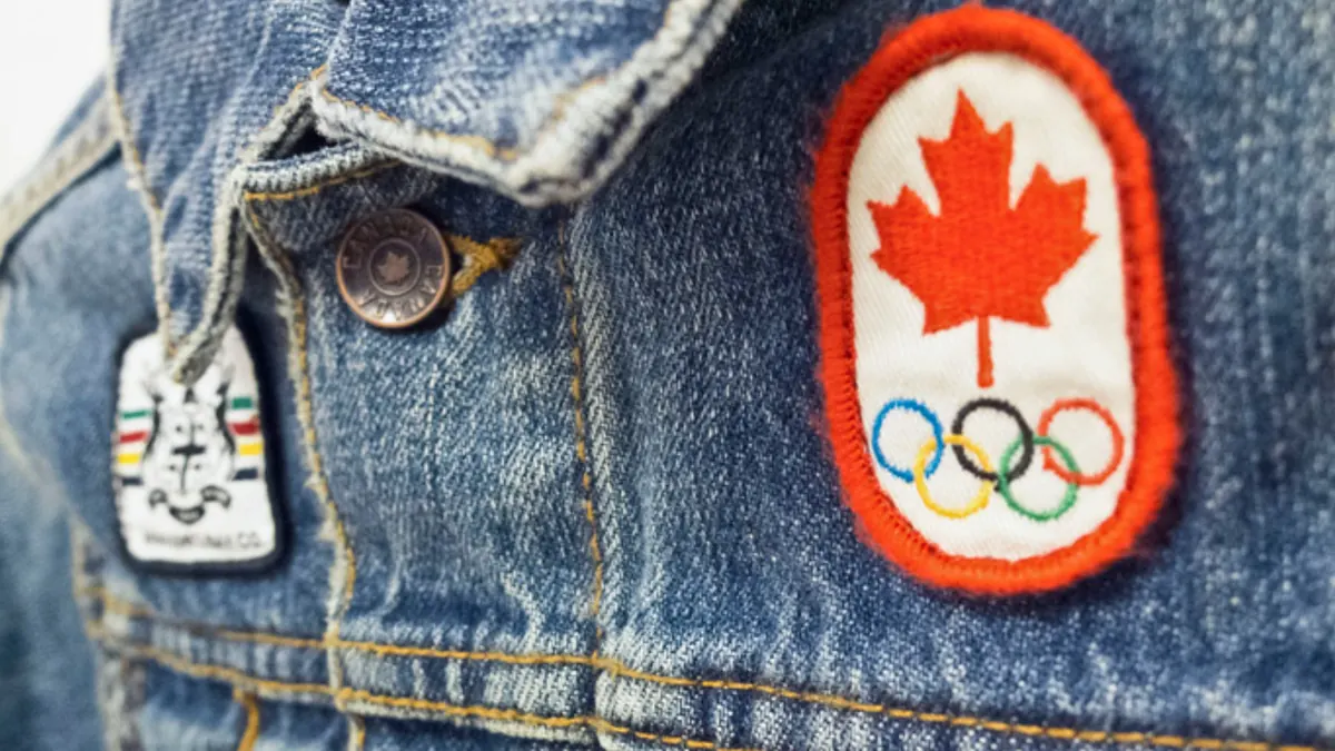 A vintage Canadian Olympic crest patch sewn onto a denim jacket beside an enamel pin.