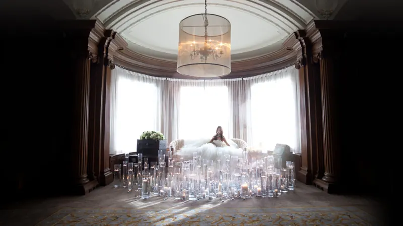 A woman in a white dress sits among hundreds of candles under a crystal chandelier and tall curtained windows in the club’s ballroom.